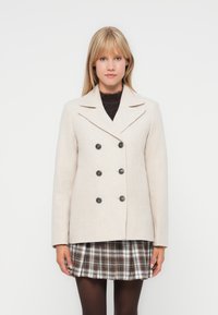 Manteau beige croisé avec des boutons noirs, col à revers et poches ; texture semblable à la laine affichée sur une jupe à carreaux.