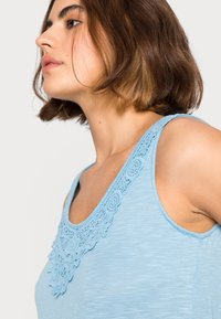 Top sem mangas azul claro com um detalhe de renda texturizada à volta do decote, um corte descontraído e uma textura de tecido suave.