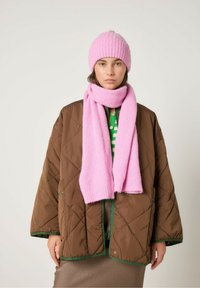 Pruun õmmeldud jakk roheliste detailidega, millele on lisatud roosa ribakudumist beanie ja sobiv sall. Mudel kannab mustrilist rohelist toppi.