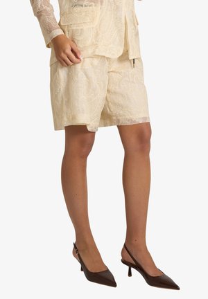 Gambe con pantaloncini in pizzo color crema e top abbinato, abbinati a décolleté slingback marrone scuro con tacco basso a gattino.