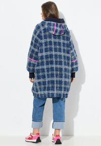 Cappotto in pile a quadri blu con cappuccio, caratterizzato da dettagli rosa. Taglio morbido con maniche lunghe, abbinato a pantaloni in denim arrotolati e scarpe da ginnastica rosa.