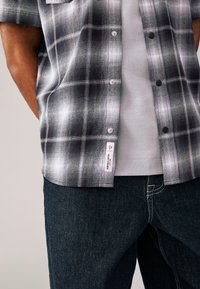 Kortärmad plaidskjorta i svart och vit flanell över en grå T-shirt, kombinerad med mörka denimjeans med kontrasterande sömmar.