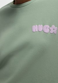 Světle zelená mikina s vyvýšenými růžovými písmeny "NUGA" a květinou na prsou, kterou nosí �člověk.