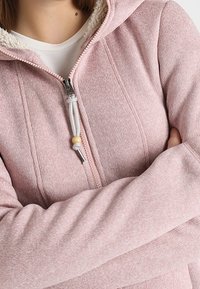 Felpa rosa con zip, caratterizzata da un rivestimento morbido e peloso, cuciture verticali e cordini con dettagli in legno. Il tessuto ha un aspetto testurizzato.