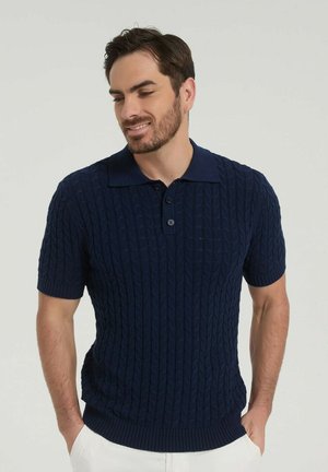 Homme aux cheveux courts foncés et à la barbe, portant un polo bleu marine en maille torsadée à manches courtes et un pantalon blanc, les mains dans les poches, souriant vers le bas.