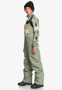Groene geïsoleerde sneeuwoverall met verstelbare schouderbanden, een voorzak met een oranje accent, gecombineerd met een gestreept lange mouw top en een zwarte beanie.