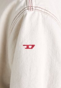 Witte stof met zichtbare textuur, met een rood geborduurd logo op de mouw. De gestikte details langs de naad zijn prominent.