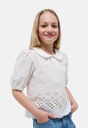 Fille souriante aux cheveux blonds raides, portant un chemisier blanc à manches bouffantes et broderie florale en œillet, les mains dans les poches d'un jean bleu.