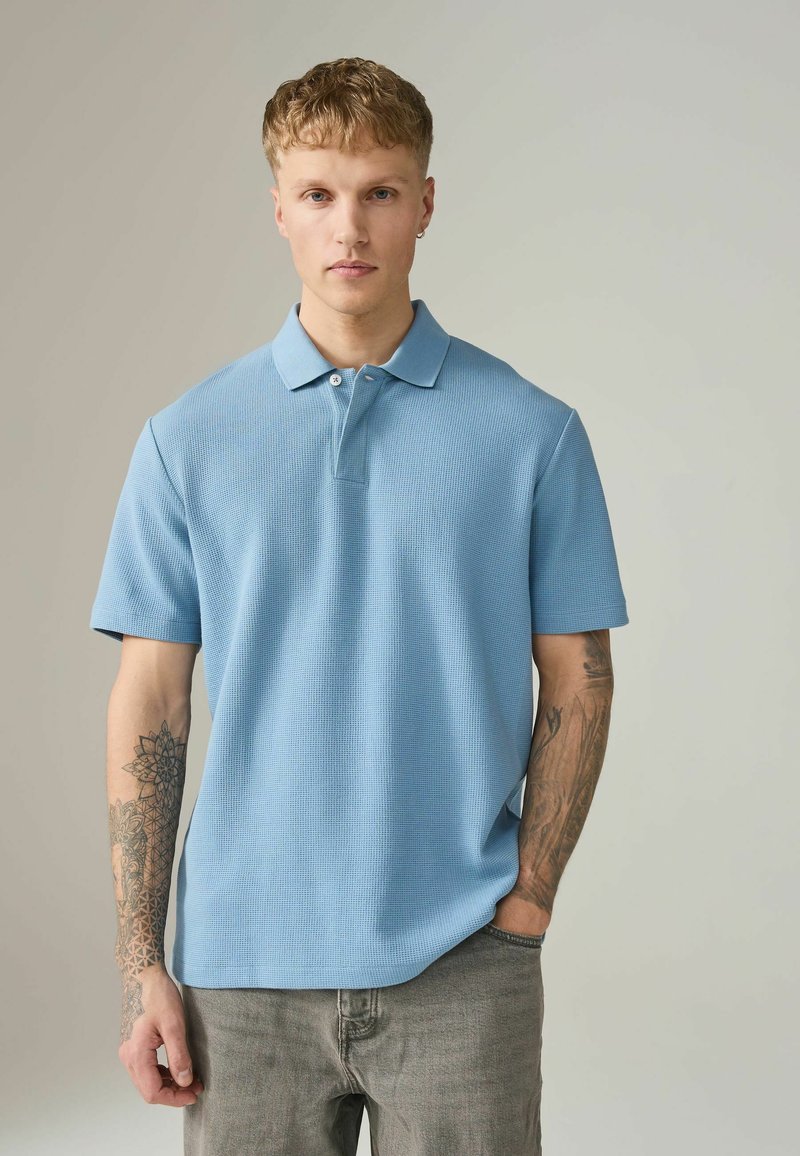 Polo bleu clair à manches courtes, tissu texturé, patte de boutonnage à deux boutons et col classique. Jean gris et tatouage visible.