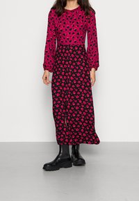 Lång klänning med en ljusrosa överdel med svarta hjärtan, som kontrasterar med en svart kjol prydd med röda hjärtan. Svarta ankelstövlar fulländar looken.