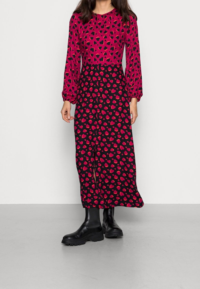 Lång klänning med en ljusrosa överdel med svarta hjärtan, som kontrasterar med en svart kjol prydd med röda hjärtan. Svarta ankelstövlar fulländar looken.