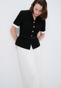 Schwarzer Blazer mit kurzen Ärmeln und goldenen Knöpfen, mit weißen Akzenten an den Ärmelbündchen und einem passenden schwarzen Gürtel; kombiniert mit einem weißen Faltenrock.