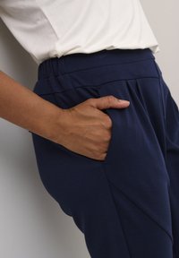 Pantalone od mekanog materijala u tamnoplavoj boji s elastičnim pojasom, bočnim džepovima i nabranim dizajnom. Minimalistički stil i glatka tekstura.