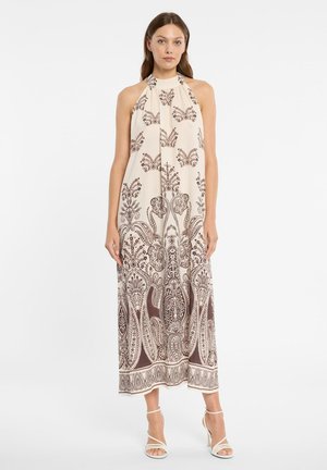 Frau trägt ärmelloses, cremefarbenes Maxikleid mit braunen, filigranen Blumen- und Paisleymustern, kombiniert mit weißen, Riemchen-High Heels, steht aufrecht.