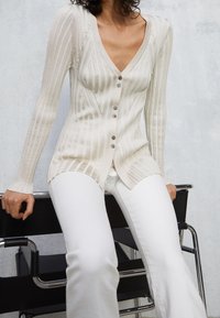 Lichtbeige geribde cardigan met een diepe V-hals, vijf knopen en lange mouwen. Gepaarde met witte flared pants van een gestructureerde stof.