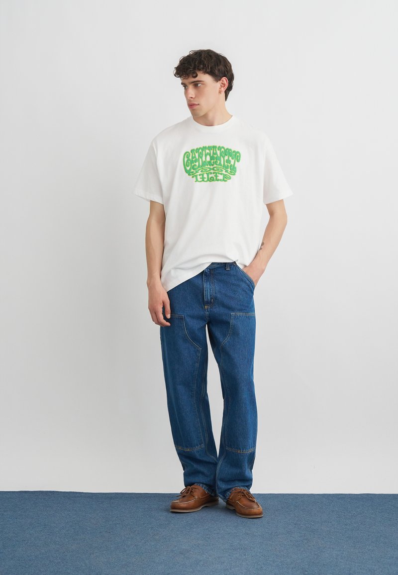 Giovane uomo che indossa una t-shirt bianca con grafica e testo verde, jeans blu con tasche applicate e scarpe marroni con lacci, in piedi contro un muro bianco.