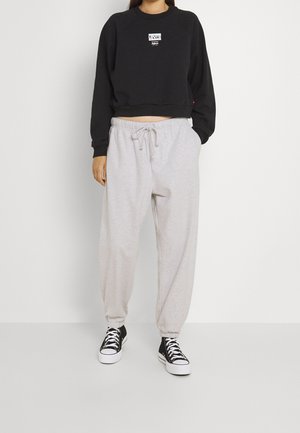 Person iført en sort Levi's cropped sweatshirt, lysegrå joggebukser med snøre og sorte højtop-sneakers, stående mod en ensartet hvid baggrund.