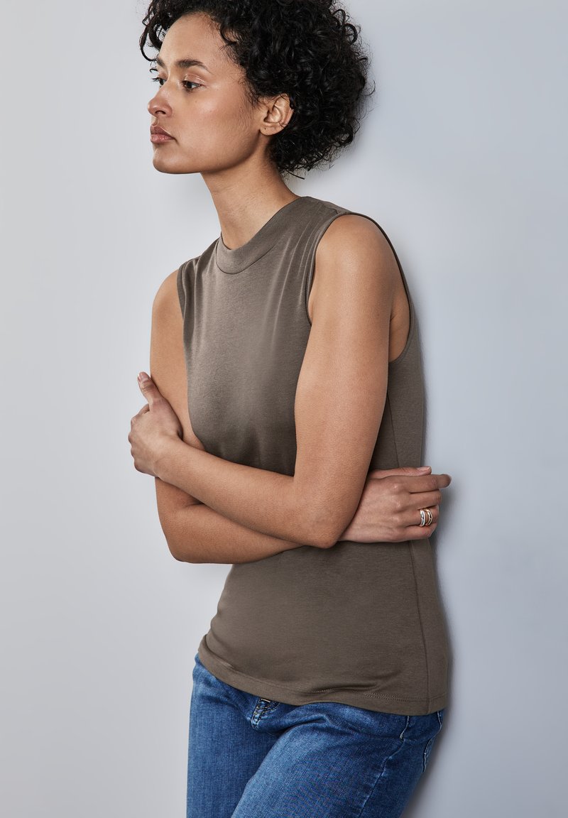 Top senza maniche, con scollatura tonda in verde oliva, realizzato in un tessuto liscio. Abbinato a jeans blu, si erge su uno sfondo grigio chiaro.