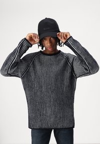 Schwarz- und grau gestreifter Strickpullover mit Oversized-Passform; Rundhals und gerippte Bündchen. Trägt eine schwarze Mütze mit „BOSS“-Logo auf der Vorderseite.