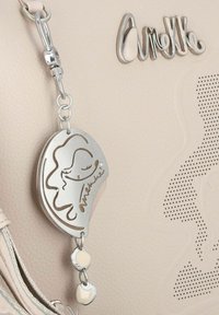 Beige Handtasche mit Textur, die einen silbernen Akzentanhänger mit ausgeschnittenem Gesichtsmotiv und dem Namen "Aneke" zeigt, befestigt mit einer Metallkette.