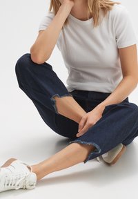 Maglietta bianca a costine con maniche corte abbinata a jeans scuri in denim con orli sfrangiati. Calzature: sneakers bianche con dettagli marroni.