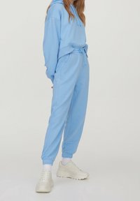 Helles blaues Hoodie mit Kordelzügen, kombiniert mit passenden schmalen Jogginghosen. Beide aus weichem, glattem Stoff. Weiße Sneaker runden das Outfit ab.