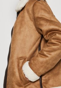 Giacca in shearling marrone con colletto in pelliccia sintetica, polsini a costine e una superficie liscia. Presenta tasche laterali e una chiusura a zip resistente.