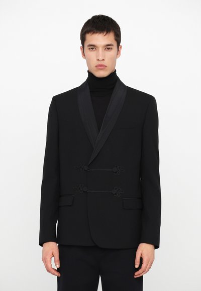 Schwarzer Blazer mit satinierter Schalkragen. Verfügt über einen dekorativen schwarzen Verschluss mit floralen Details und seitlichen Taschen. Getragen über einem schwarzen Rollkragenpulli.