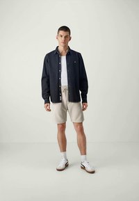Young man stands in casual outfit wearing a black button-up shirt over a white tank top, beige shorts, white socks, and white sneakers.