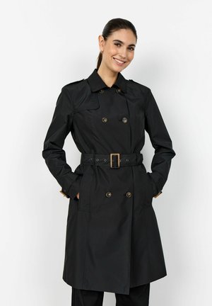 Femeie zâmbitoare, purtând un palton trench negru cu două rânduri de nasturi și curea, cu mâinile în buzunare, pe un fundal alb simplu.
