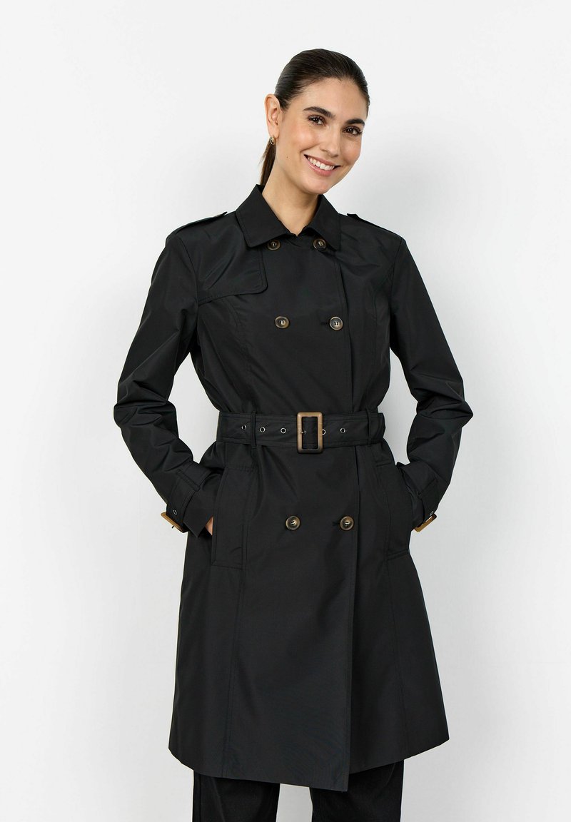 Frau lächelnd, trägt einen schwarzen, doppelt geknöpften Trenchcoat mit Gürtel, die Hände in den Taschen, vor einem schlichten weißen Hintergrund.