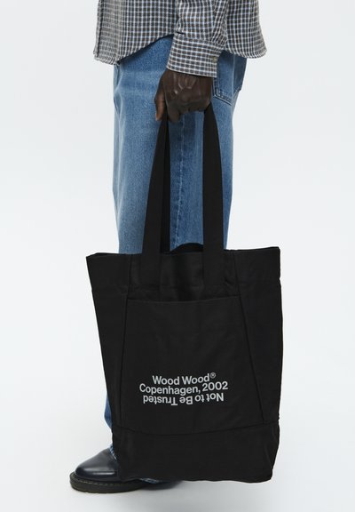 Fekete vászontáska két füllel, fehér nyomtatott szöveggel. Egyszerű dizájn, téglalap alak, és elülső zsebének részlete.