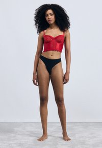 Top corto in pizzo rosso con coppe a stecche e orlo scallopato abbinato a perizoma nero. Texture liscia, design aderente e sp straps regolabili.