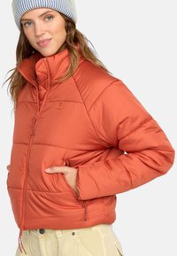 Oranje pufferjack met een hoge kraag, ritssluiting aan de voorkant en zijzakken. Beschikt over gestikte details en een klein logo-opdracht op de borst.