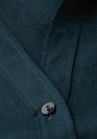 Camisa de lino azul oscuro con un botón negro, costuras visibles y una textura suave. El collar se superpone con un acabado limpio.