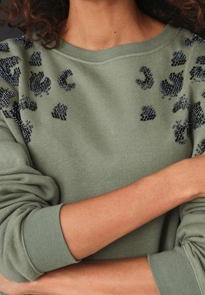 Personne portant un pull vert à col rond avec des détails léopard en perles noires, les bras croisés sur la poitrine.