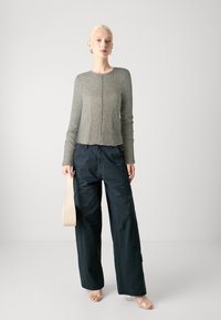 Sivi rebrasti dugi rukav skraćeni pulover u kombinaciji s širokim tamnim hlačama. Model drži svijetlosmeđi kutak. Minimalistički stil, obična pozadina.