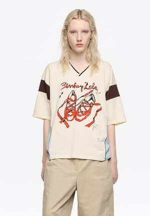 Camiseta de gran tamaño color beige con escote en V negro, que presenta una ilustración roja de zapatillas de ballet. Mangas cortas con una franja marrón y dobladillo redondeado.