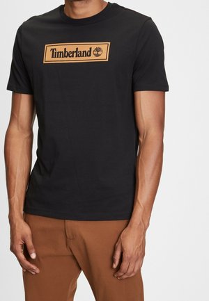 Mężczyzna w czarnej koszulce Timberland z beżowym logo na piersi i brązowych spodniach, stojący z rękami swobodnie opuszczonymi wzdłuż ciała.