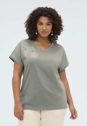 Žena s kudrnatými vlasy nosí šedé tričko s výstřihem do V a květinovým potiskem na rameni a světle zbarvené kalhoty, stojící na jednobarevném pozadí.