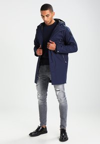 Giacca impermeabile blu navy con cappuccio, tasche con zip e tessuto testurizzato, abbinata a jeans grigi chiari strappati e scarpe nere.