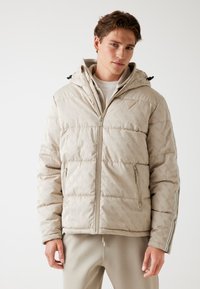 Jachetă puffer bej cu un model subtil, fermoar frontal, buzunare laterale și glugă detașabilă. Prezintă dungi albe pe mâneci.