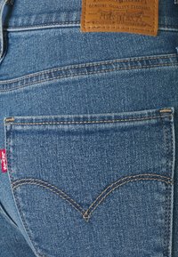 Nära bild av en bakficka på blå denimjeans med synlig orange söm, röd Levi's-tag och lädermärke ovanför fickan.