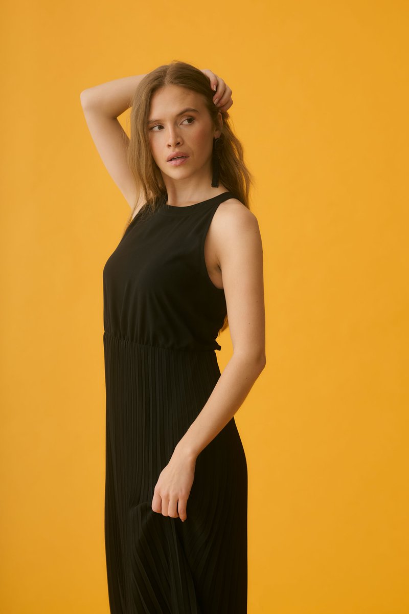Jeune femme en longue robe noire sans manches pose avec une main sur la tête et l'autre tenant la robe devant un fond jaune uni.