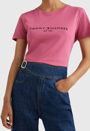 Kvinde iført lyserød Tommy Hilfiger t-shirt og blåt højtaljede jeans med bælte, den ene hånd i lommen mod ensfarvet baggrund.