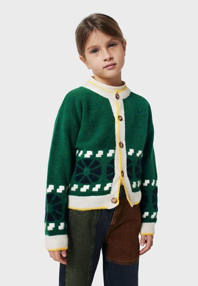 Kind, das eine grüne Strickjacke mit weißen und dunkelblauen geometrischen Mustern sowie eine Patchworkhose in Grün, Braun und Marineblau trägt.