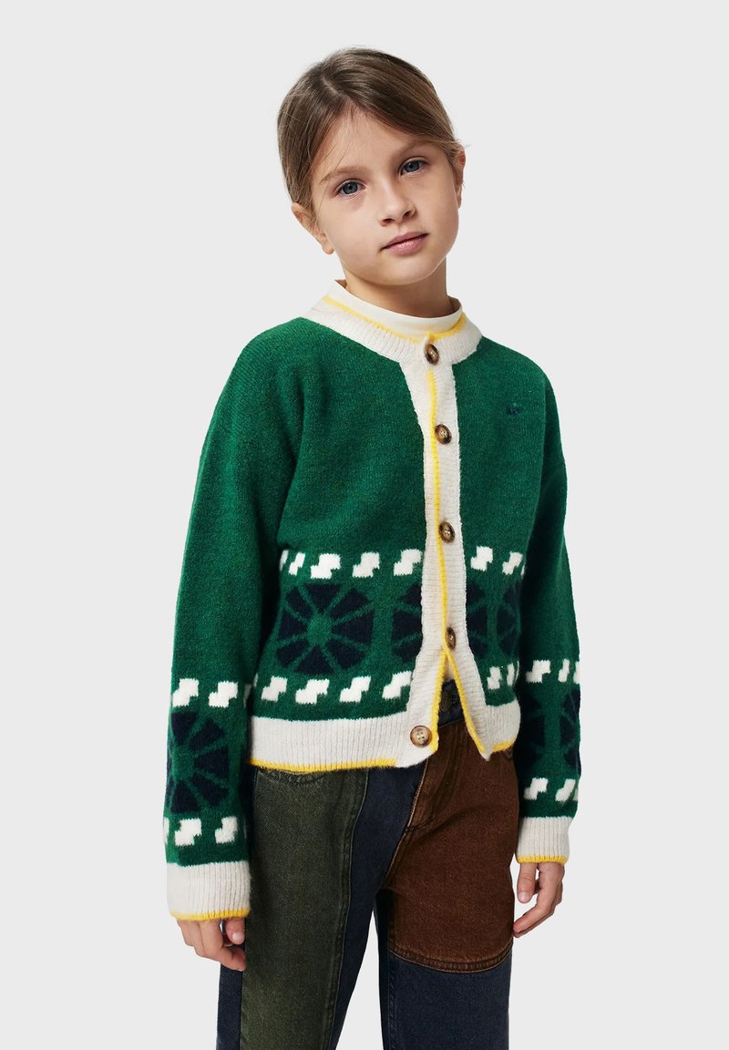 Grön ullcardigan med vit krage, gula detaljer och träknappar. Har geometriska mönster vid fållen och ärmarna.