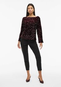 Burgunderfarbener, mit Pailletten besetzter Langarm-Top mit Bootsausschnitt, kombiniert mit schwarzen taillierten Hosen und burgunderfarbenen High Heels. Glatte Textur.