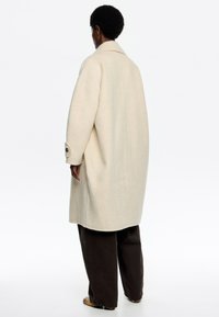 Cappotto oversize beige in lana con colletto a punta e polsini abbottonati, abbinato a pantaloni larghi marroni scuri. Tessuto morbido e silhouette rilassata.