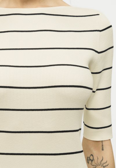 Κρέμ ριμπ πλεκτό τοπ με μαύρες οριζόντιες ρίγες και μανίκια μέχρι τον αγκώνα σε άτομο με τατουάζ στο αντιβράχιο.